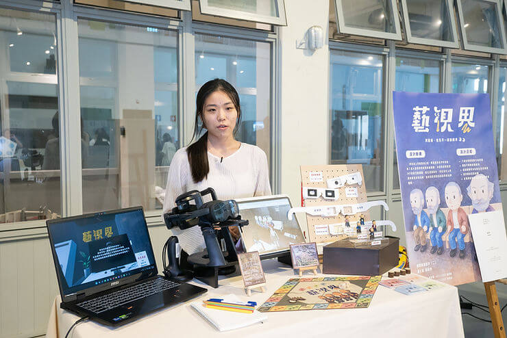 A woman stands by a table with tech gadgets, a laptop, and a poster in Chinese. Bright setting with colorful materials and a calm mood.