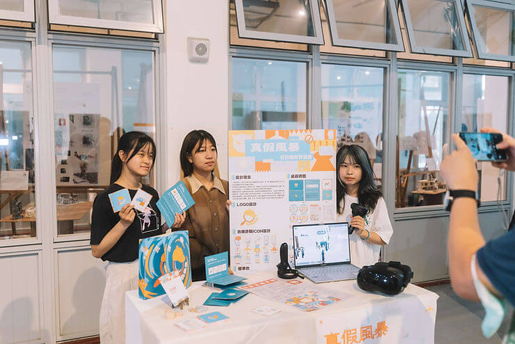 Three people present tech designs at a booth with colorful posters and VR gear. A fourth person photographs them. Bright, professional setting.